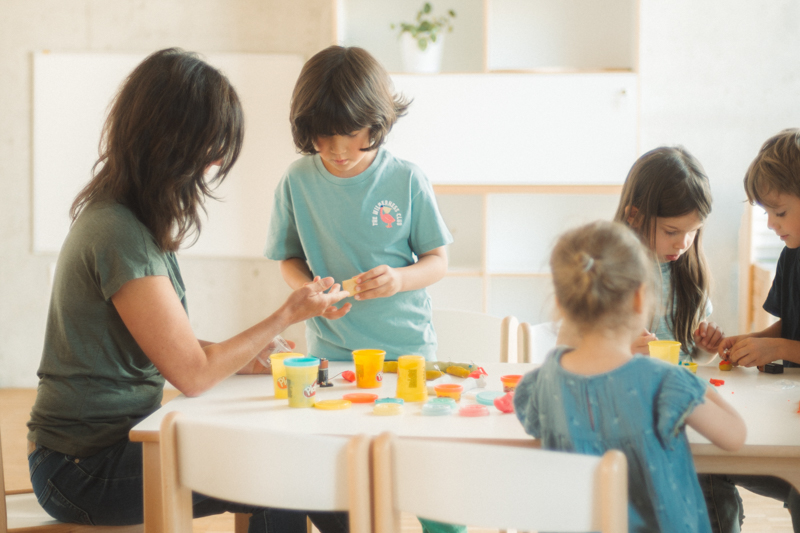 Eine Gruppe von Kindern arbeitet an einem Tisch zusammen mit Knete. Eine Erzieherin hilft dabei.