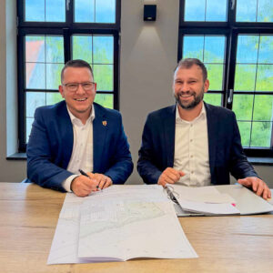 Bürgermeister von Merchweiler und Geschäftsführer von littlebigFuture sitzen zusammen vor dem unterschrieben Bauantrag und lachen zusammen in die Kamera.