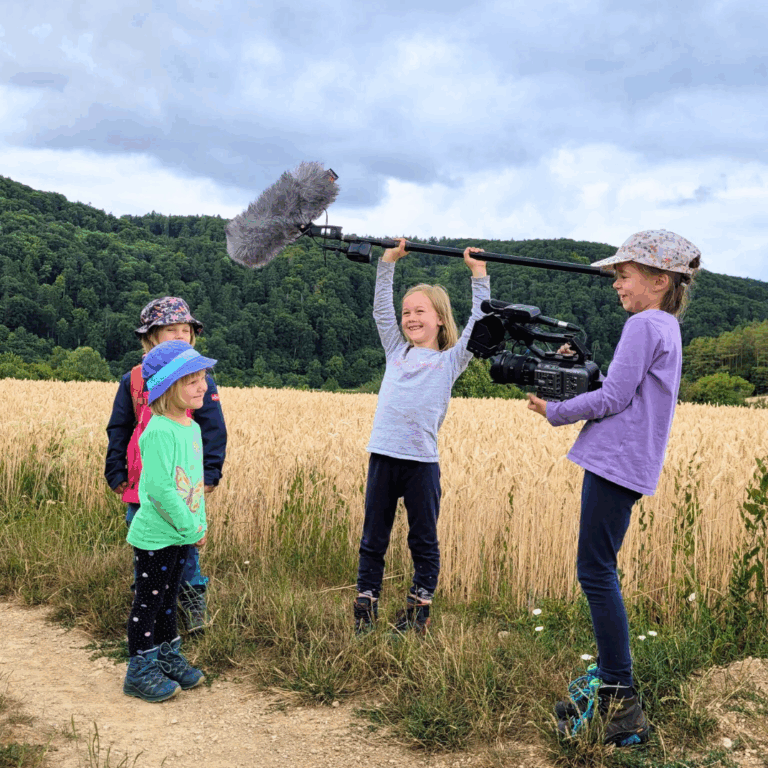 Vier Kinder in der Natur. Eins hat eine Kamera in der Hand und filmt zwei der Kinder. Das Vierte hält eine große Tonangel in beiden Händen.