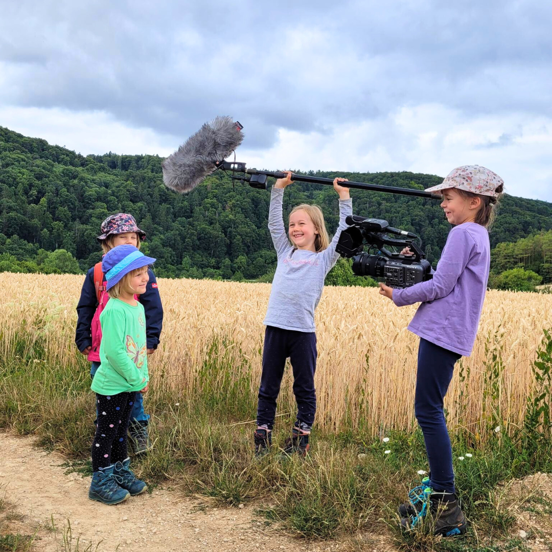 Vier Kinder in der Natur. Eins hat eine Kamera in der Hand und filmt zwei der Kinder. Das Vierte hält eine große Tonangel in beiden Händen.