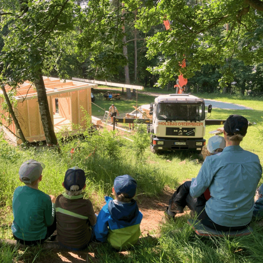 Im Hintergrund wird eine Holzhütte mit einem Kran aufgebaut. Im Vordergrund sitzen Kinder und eine Erzieherin und beobachten das Geschehen.