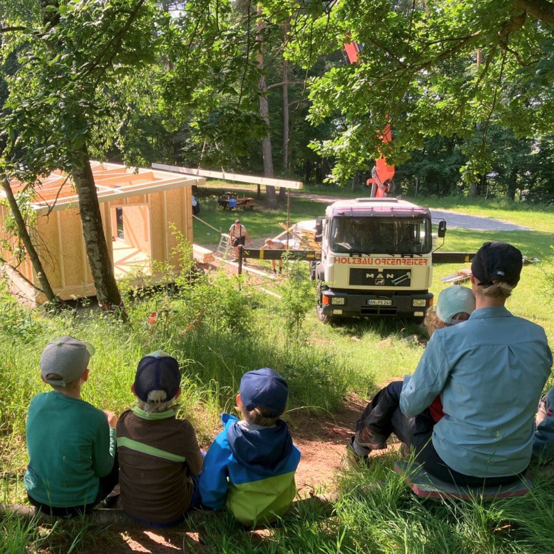Im Hintergrund wird eine Holzhütte mit einem Kran aufgebaut. Im Vordergrund sitzen Kinder und eine Erzieherin und beobachten das Geschehen.
