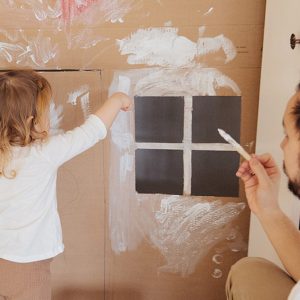 Erwachsener und Kind malen zusammen ein Haus aus Pappe an