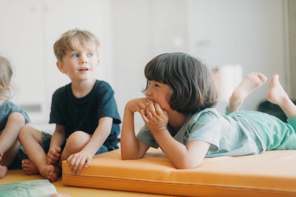 Ein Kind sitz auf einem Teppich und ein Kind liegt auf einer gelben Matte